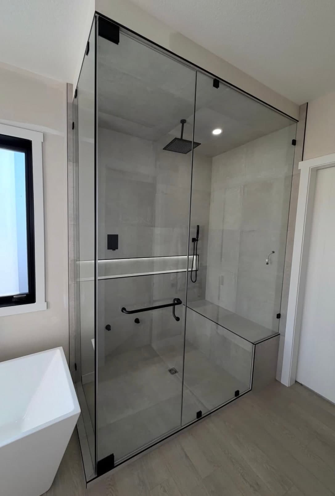 Tall glass shower with black clamps and handle, a ceiling rain head and a tiled bench